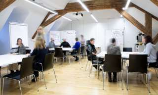Ein mittelgrosser Saal mit Dachschräge und hölzernen Dachbalken. Im Raum befinden sich mehrere beschriftete Flipcharts und Tischgruppen mit Personen, die an einem Workshop teilnehmen. 